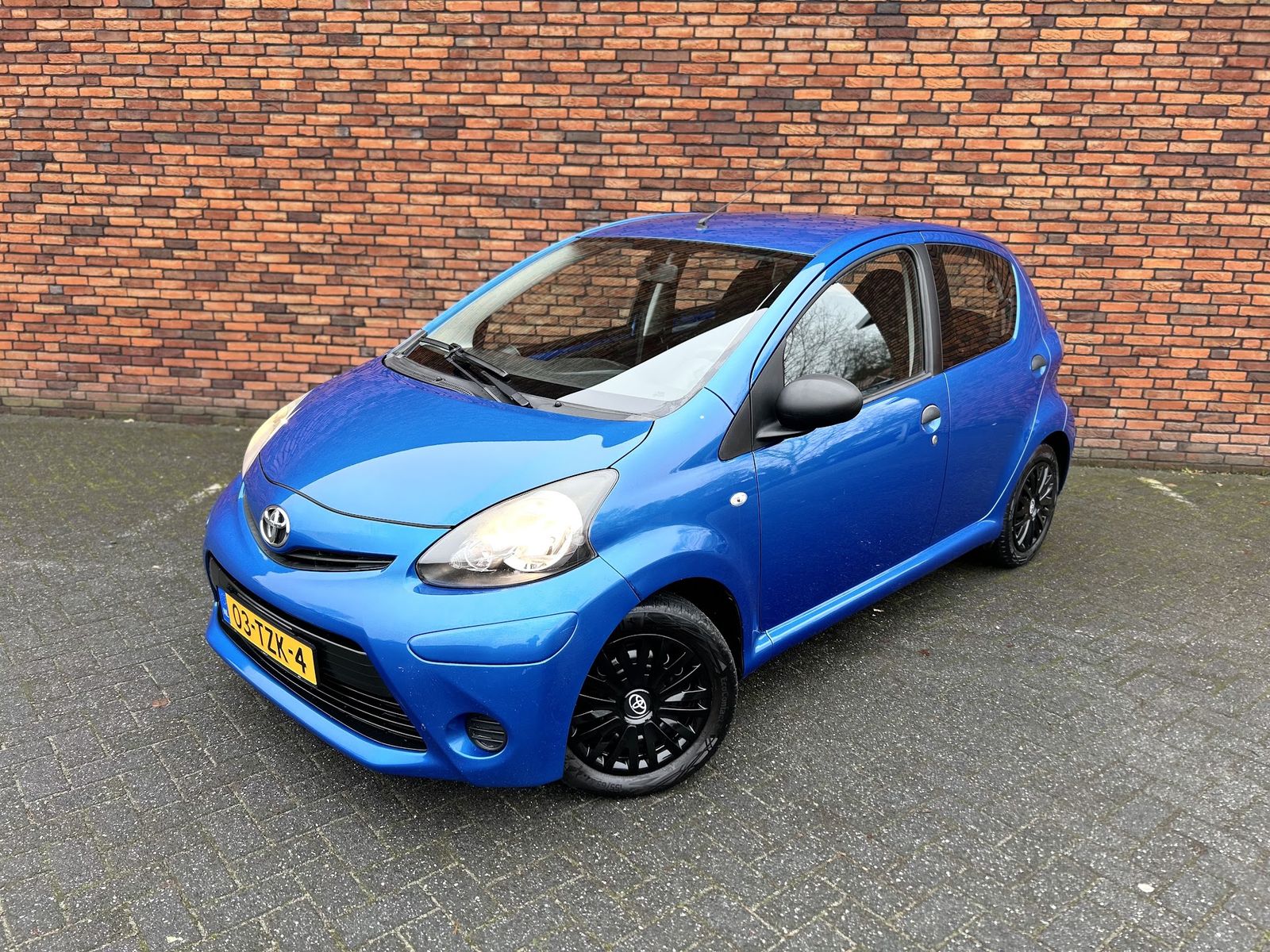 Blauwe Toyota Aygo met zwarte velgen voor de bakstenen muur